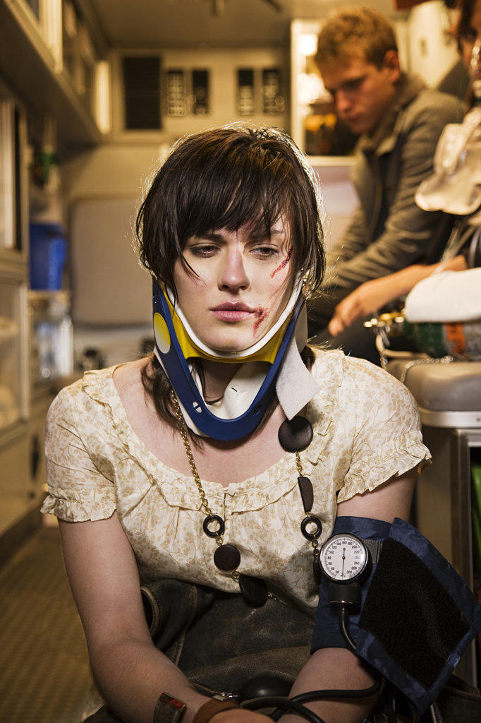 Injured woman wearing a neck brace sitting on back of open ambulance, while her friends are getting treated in the back 