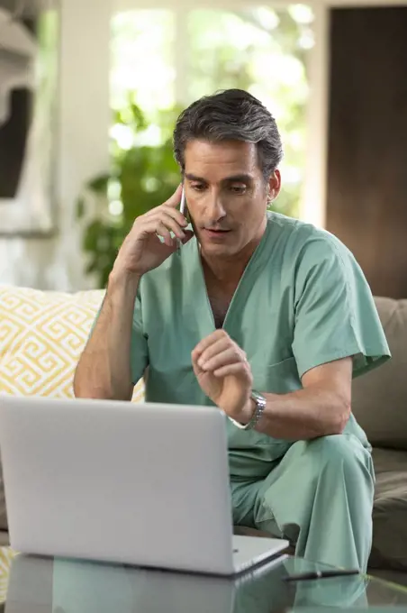 Hispanic Male doctor practicing tele-medicine from his home, using cell phone and laptop computer, Talking to patient on video call 
