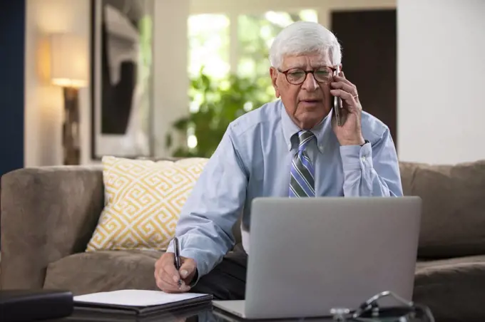 Mature Caucasian Male doctor practicing tele-medicine from his home using laptop computer, Talking to patient and taking notes