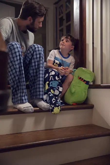 Father and son sitting at top of stairs in home getting ready for bedtime 