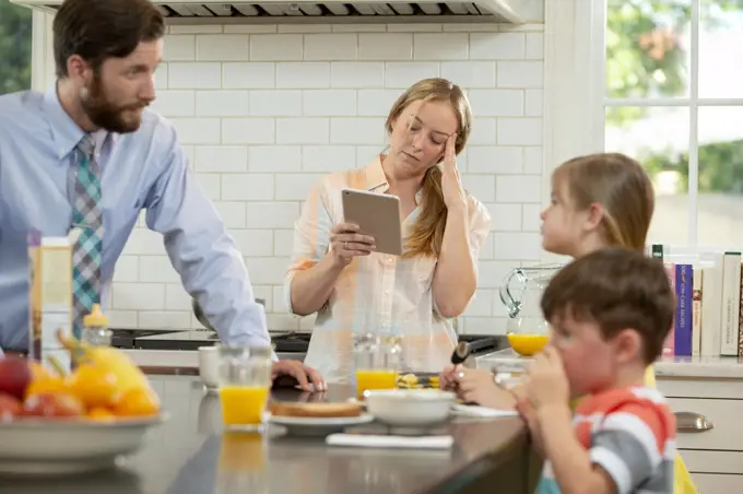 Mom with family in kitchen, struggling to get the kids breakfast and ready to start the day. Checking tablet to connect with doctor and ask about her headaches