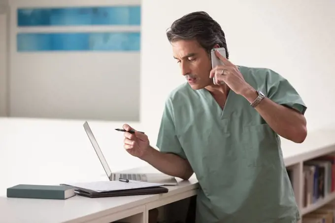 Hispanic Male doctor in his home practicing Telemedicine, talking on cell phone and taking notes 