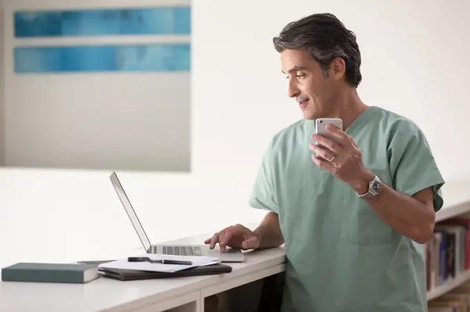 Hispanic Male doctor in his home practicing Telemedicine, talking on cell phone and using laptop computer 