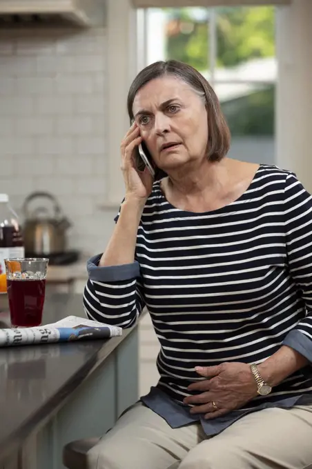 Senior Caucasian woman in her kitchen describing her abdominal pain to doctor on the phone