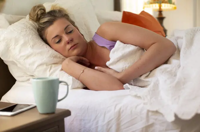 Young woman in her bed debating getting out of bed, smartphone and coffee mug sitting on bedside table 
