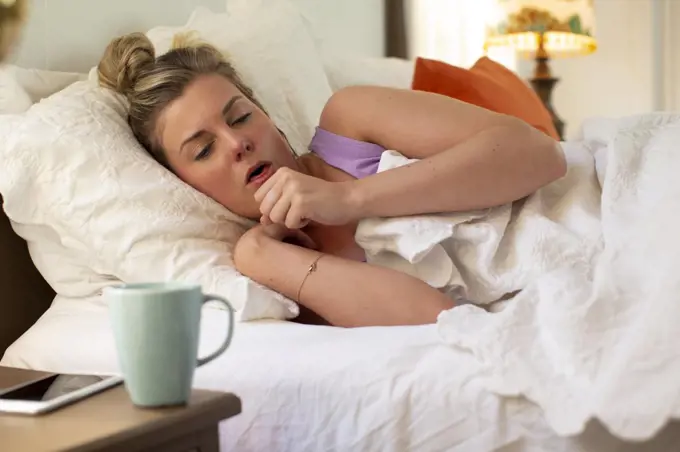 Coughing young woman in her bed debating getting out of bed, smartphone and coffee mug sitting on bedside table 