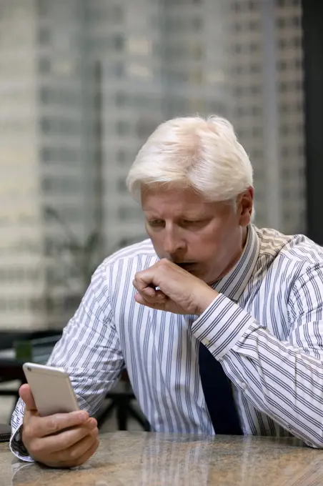 Coughing mature Caucasian man fighting off a cold in office of high-rise building downtown, looking at smartphone having a tele-medicine video call with doctor 