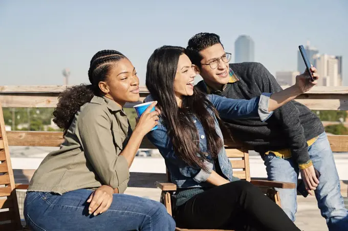 Group of young co-workers hanging out on rooftop patio, two women and man taking selfie with mobile phone 