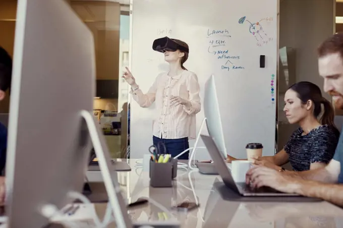 Co-Workers working on computer in office while woman is using Virtual Reality Headset standing in front of dry erase board with flowchart in background 