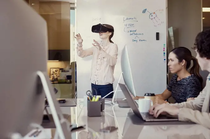 Co-Workers working on computer in office while woman is using Virtual Reality Headset standing in front of dry erase board with flowchart in background 