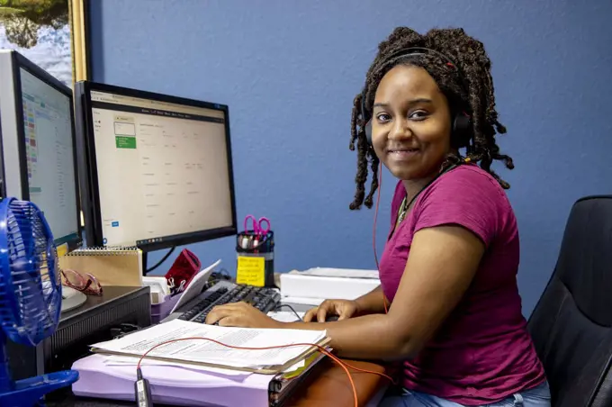 A portrait of an African American woman at her call center desk and her headset on, working from home, looking into camera smiling.