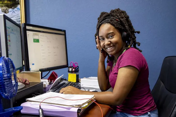 A portrait of an African American woman at her call center desk and her headset on working from home, looking into camera smiling.