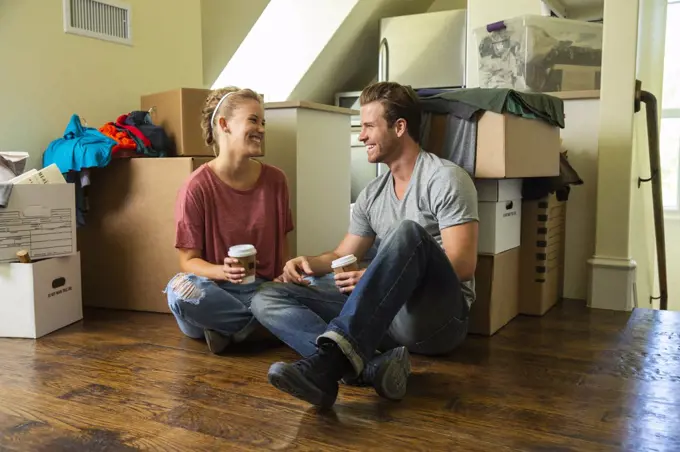 Young couple with coffee sitting on floor with boxes while moving in to new home 