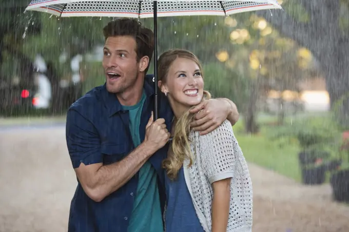 Young couple walking through park with umbrella in the rain, both looking out at the falling rain 
