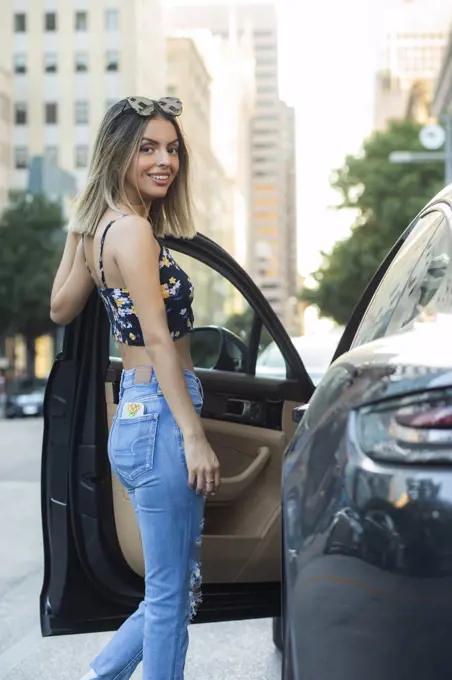 Portrait of Hispanic woman wearing floral top with torn blue jeans, getting into car on busy city street 
