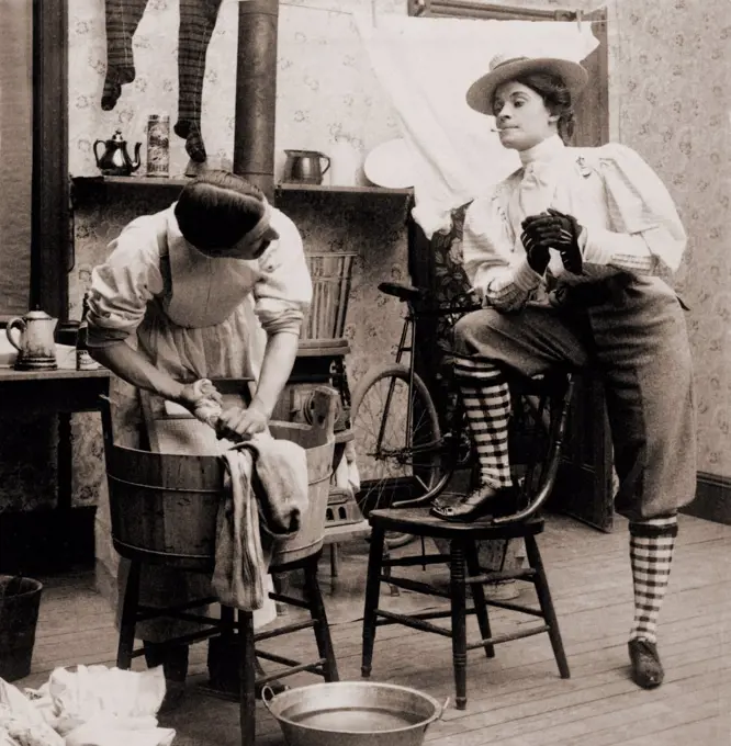 Humorous scene of life with 'the new woman.' Woman smoking and wearing knickers postures arrogantly as a man drudges over laundry. 1901.
