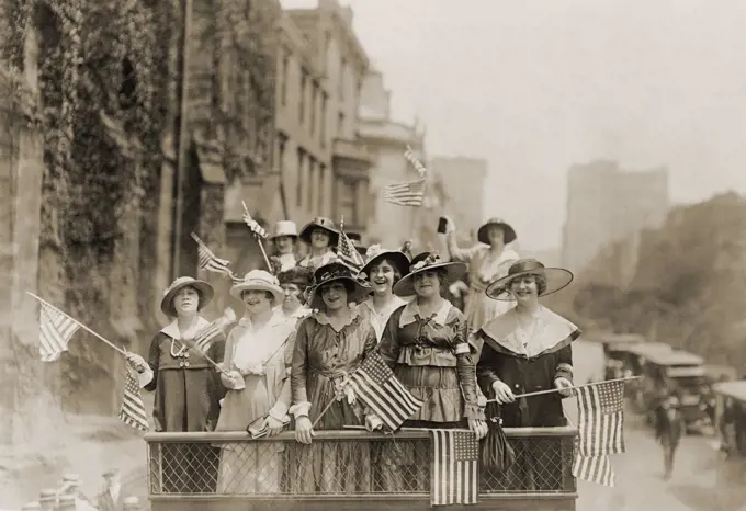 Smiling young women waving flags from the top of an open vehicle in a New York City parade. Ca. 1910-1915.