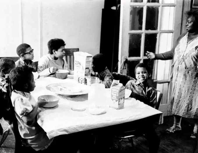 An African American mother serves six children a breakfast of corn flakes and milk in their apartment in New York's Harlem district. The large jar on the table serves as the family's sugar bowl. April 6, 1968.