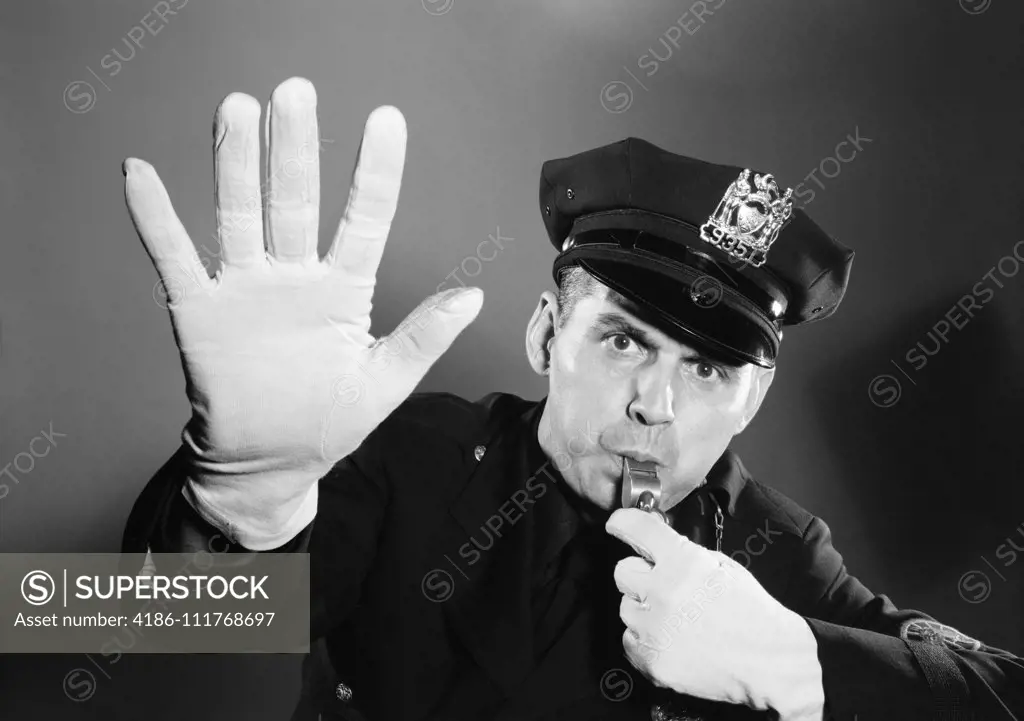 1950s 1960s POLICE MAN LOOKING AT CAMERA HAT WITH BADGE BLOWING WHISTLE WHITE GLOVED HAND UP TO HALT STOP TRAFFIC
