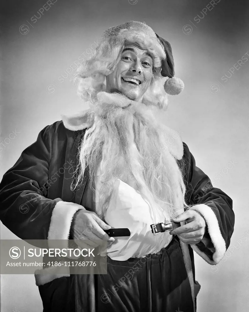 1950s SMILING MAN DRESSING UP IN SANTA COSTUME HOLDING BELT AROUND PADDED STOMACH