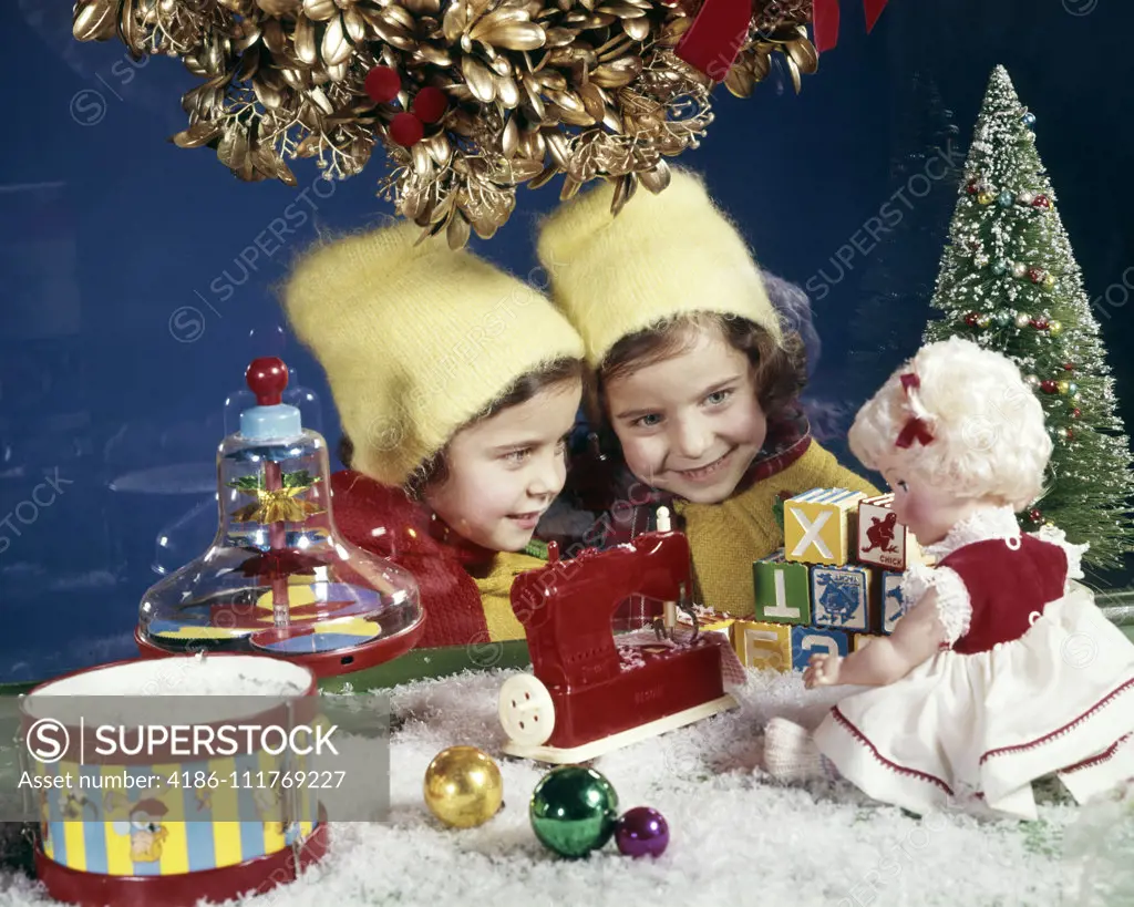 1960s TWO SMILING GIRLS TWIN SISTERS LOOKING IN CHRISTMAS TOY STORE WINDOW AT DOLL AND SEWING MACHINE