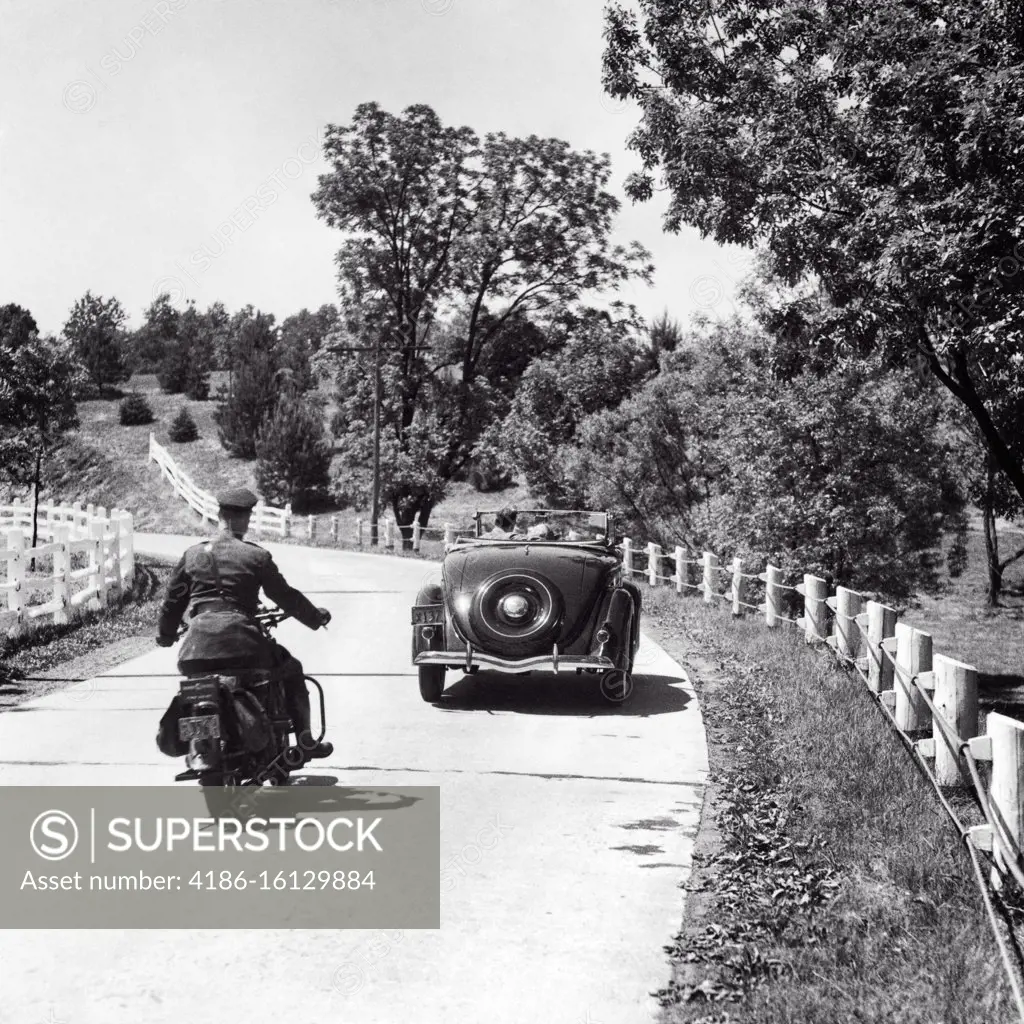 1930s 1940s MAN HIGHWAY PATROL POLICE OFFICER ON MOTORCYCLE PURSUING CONVERTIBLE CAR WITH COUPLE TWO PASSENGERS ON COUNTRY ROAD