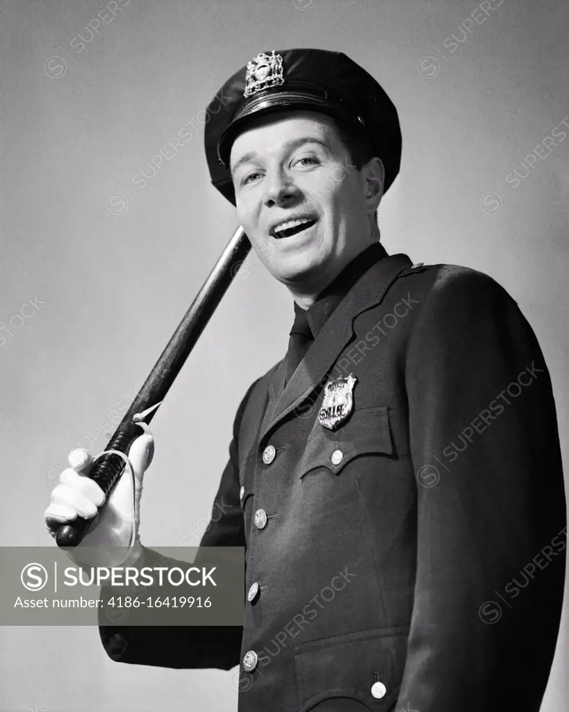 1950s SMILING UNIFORMED BEAT POLICEMAN LOOKING AT CAMERA TIPPING HIS HAT WITH NIGHT STICK BILLY CLUB