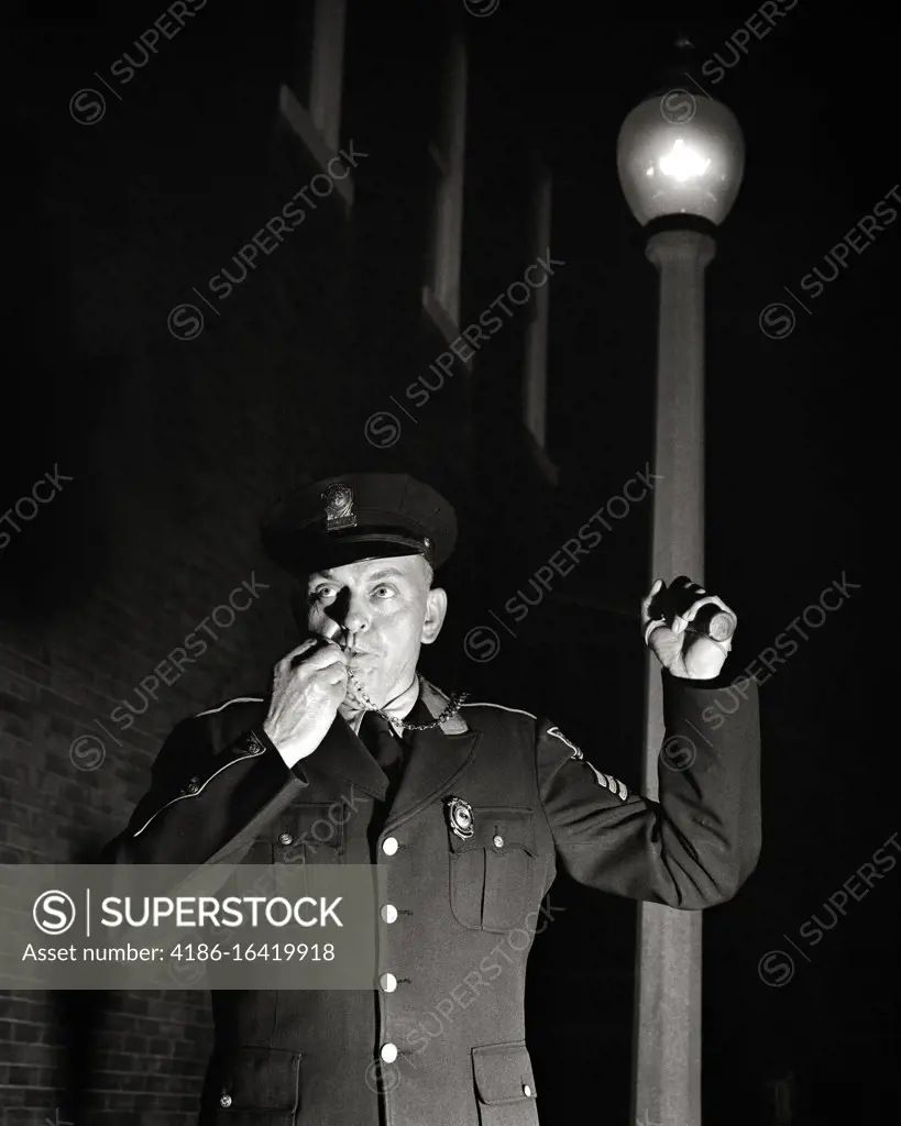 1950s SINGLE FOOT PATROL POLICE OFFICER SERGEANT IN UNIFORM AT NIGHT LOOKING AT CAMERA HOLDING FLASHLIGHT BLOWING WHISTLE