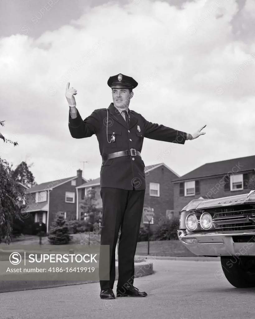 1960s SINGLE SMILING UNIFORMED POLICEMAN ARMS HANDS EXTENDED STANDING AT INTERSECTION DIRECTING SUBURBAN AUTOMOBILE TRAFFIC 