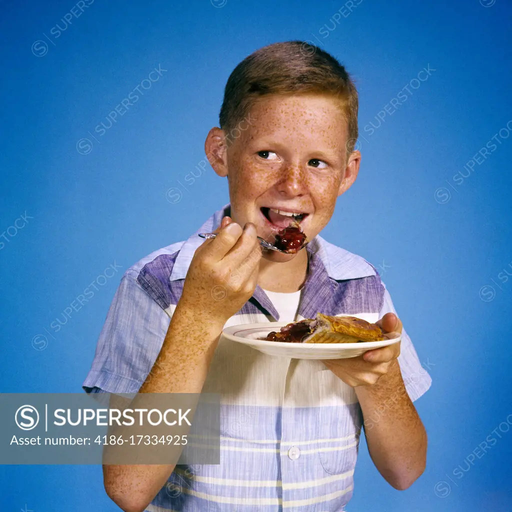 1960s 1970s SMILING FRECKLE-FACED BOY EATING ENJOYING SLICE OF BERRY CHERRY PIE LOOKING OFF CAMERA