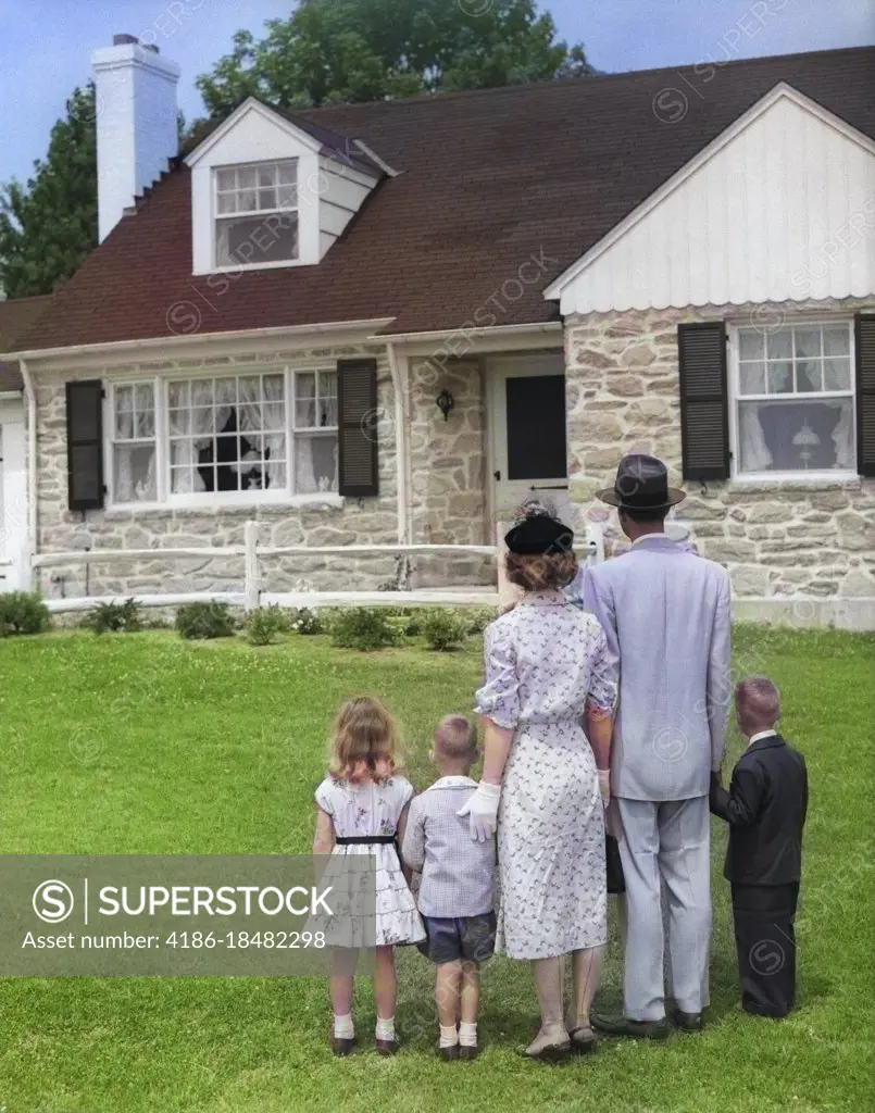 1950s FAMILY OF FIVE WITH BACKS TO CAMERA ON LAWN LOOKING AT FIELDSTONE SUBURBAN HOUSE