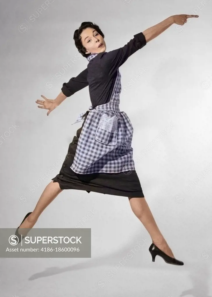 1950s WOMAN HOUSEWIFE IN GINGHAM APRON RUNNING AND JUMPING INTO AIR SMILING AND POINTING INDEX FINGER LOOKING AT CAMERA