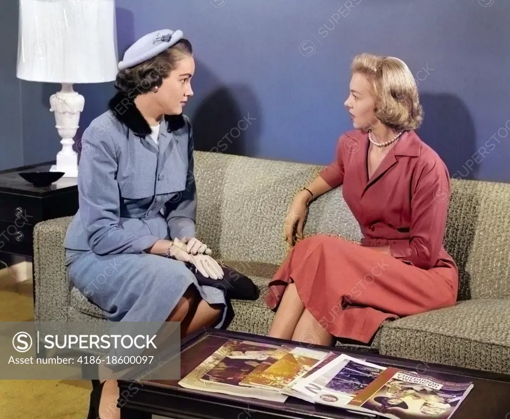 1950s PAIR OF WOMEN SITTING ON LIVING ROOM SOFA TALKING ONE WEARING SUIT WITH HAT & GLOVES