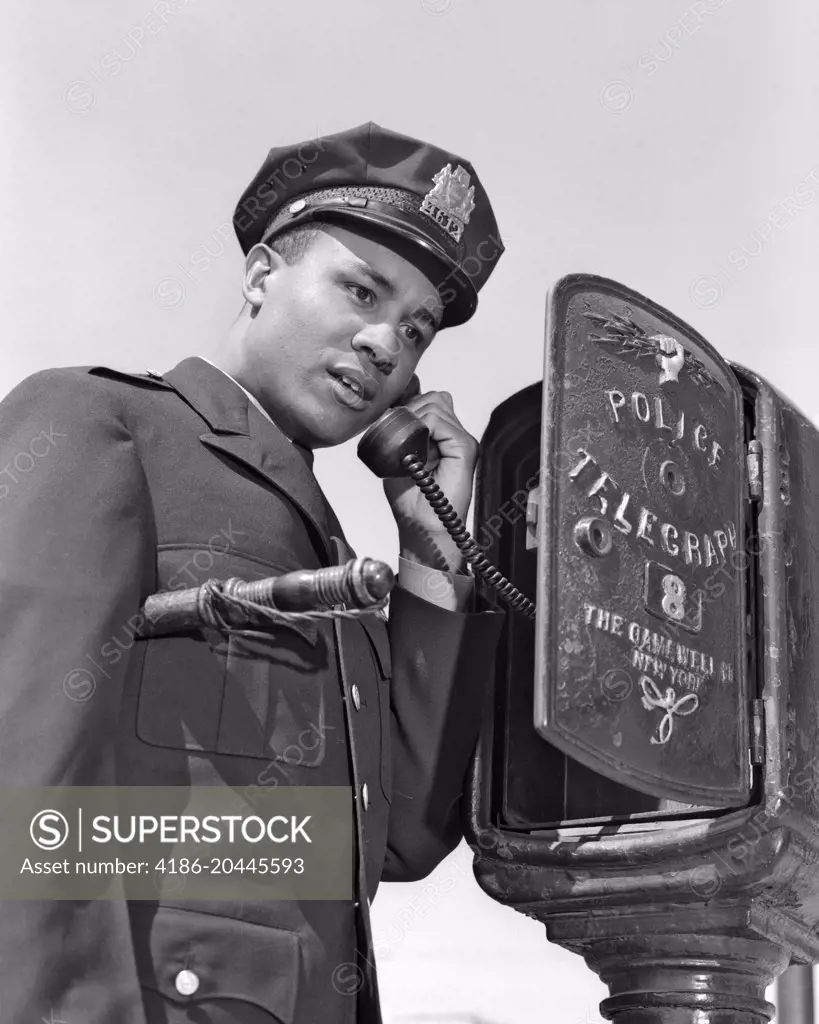 1960s AFRICAN AMERICAN POLICE OFFICER MAKING CALL AT POLICE BOX TELEPHONE NIGHT STICK TUCKED UNDER HIS ARM