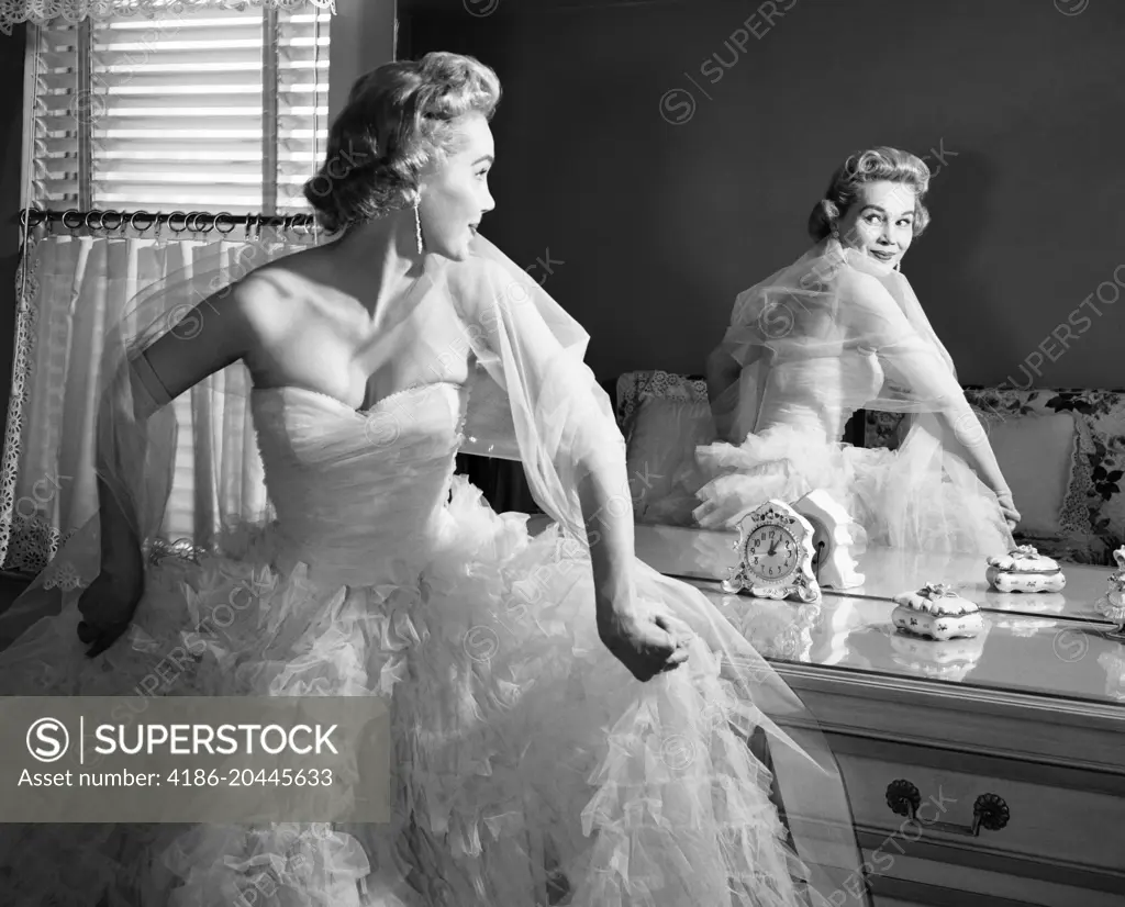 1950s BLONDE IN STRAPLESS GOWN WITH CRINOLINES TURNING TO LOOK AT BACK VIEW OF SELF IN MIRROR