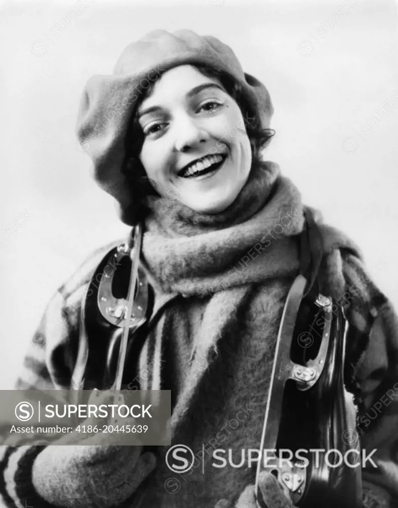 1920s 1930s SMILING WOMAN DRESSED FOR WINTER LOOKING AT CAMERA PAIR OF ICE SKATES OVER HER SHOULDERS