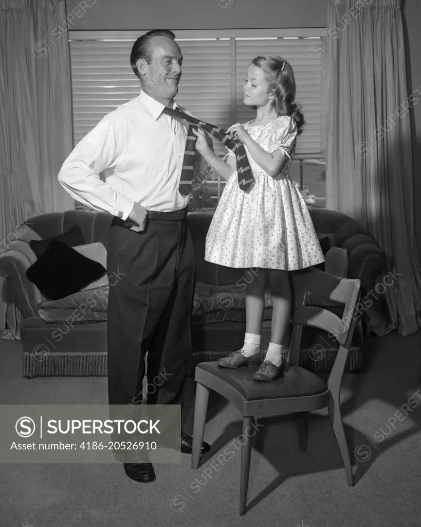 1950s CONFIDENT LITTLE GIRL STANDING ON CHAIR TYING NECKTIE FOR HER SMILING FATHER