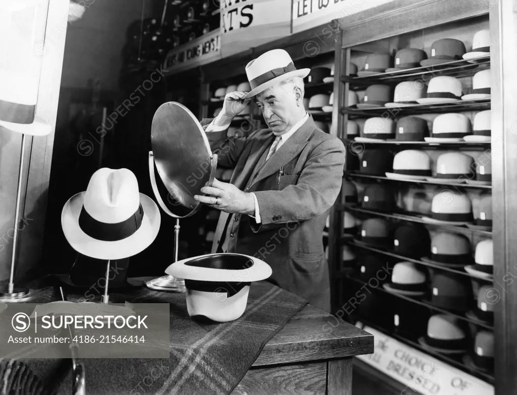 1940s SENIOR MAN TRYING ON HATS LOOKING IN MIRROR IN HAT STORE