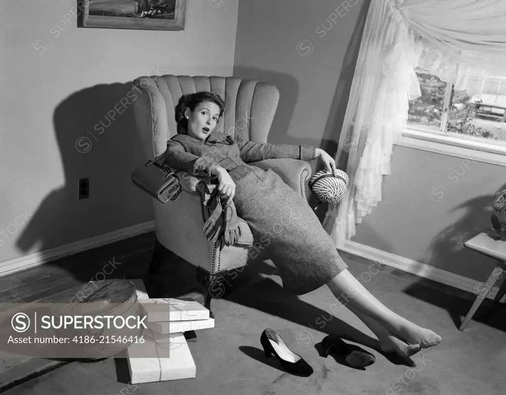1950s WOMAN EXHAUSTED FROM SHOPPING SLUMPING IN LIVING ROOM CHAIR SHOES OFF PACKAGES EVERYWHERE LOOKING AT CAMERA 