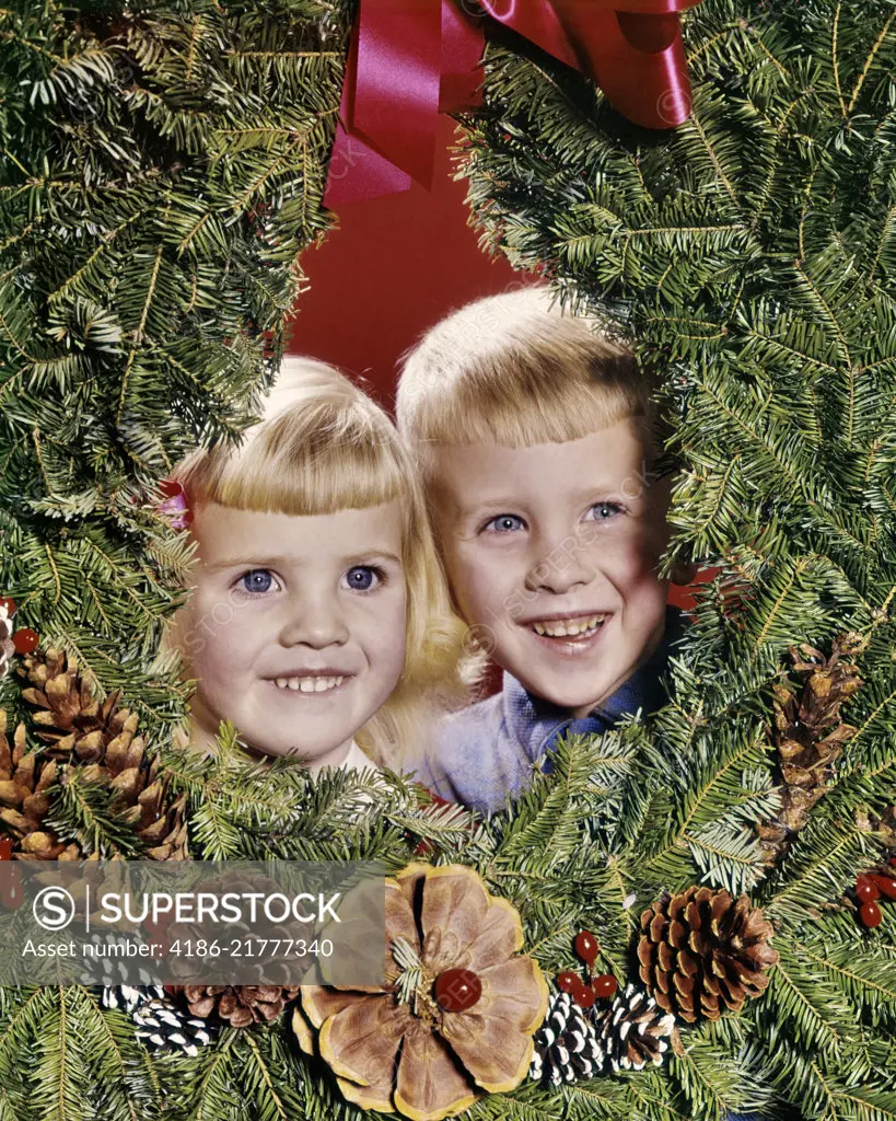 1960s SMILING BLONDE BOY AND GIRL BROTHER AND SISTER PEEKING OUT FROM CENTER OF CHRISTMAS WREATH