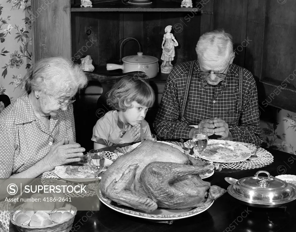 1950S 1960S Little Girl Look At Turkey Sitting Between Grandmother Grandfather Saying Grace At Table