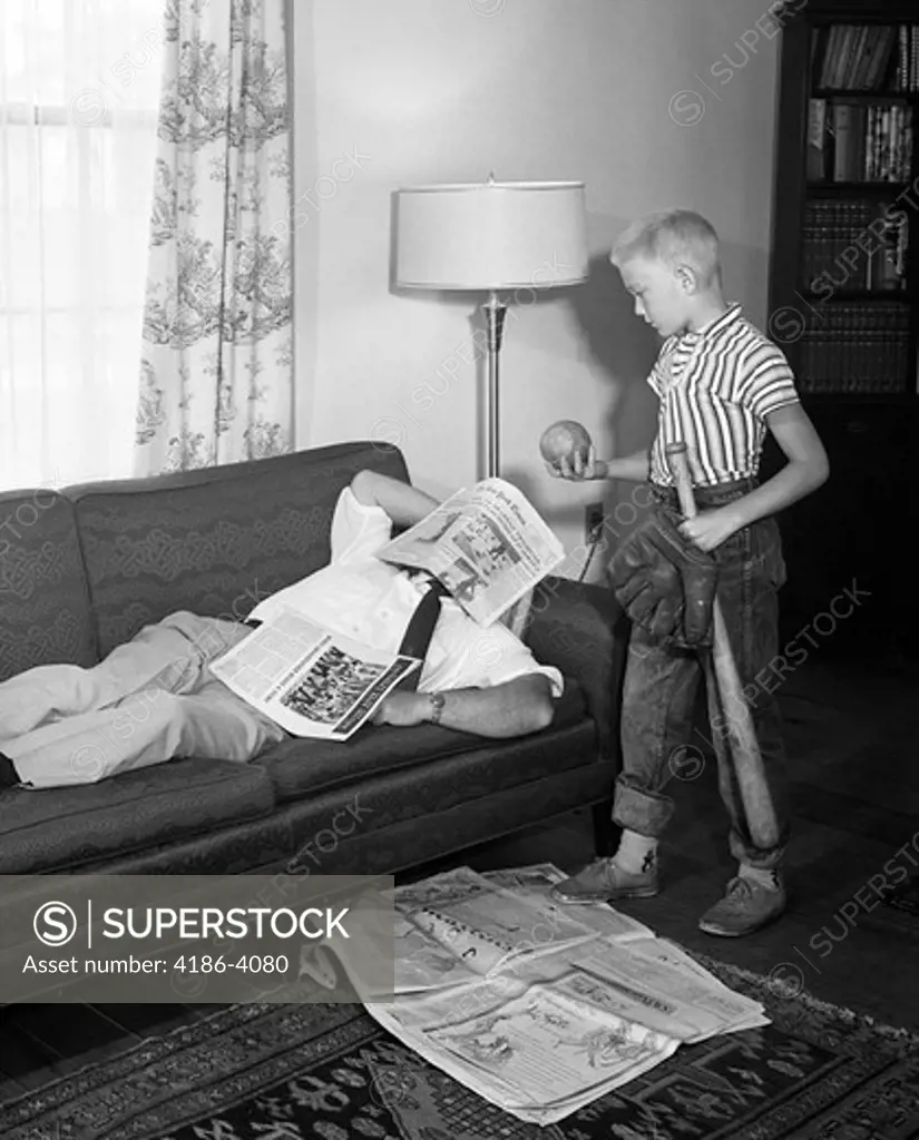 1950S Father Lying On A Sofa With Newspaper Over His Head While Son Is Standing Over Him With Bat Ball And Baseball Glove