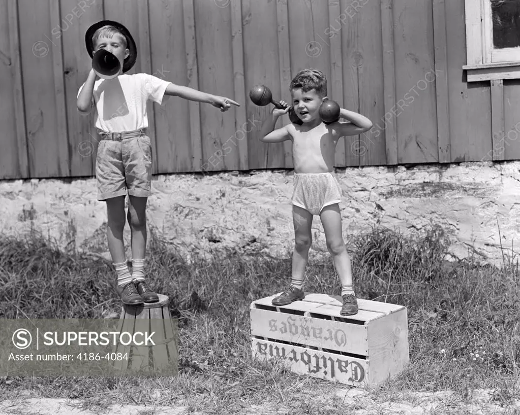 1930S 1940S Boys Playing Carnival Strongman One Lifting Dumbbells Other Announcing Act Through Megaphone