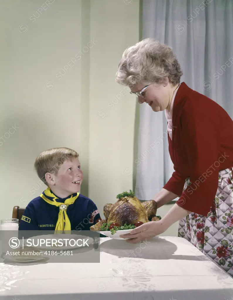 1960S Grandmother Putting Turkey On Table Near Boy In Cub Scout Uniform Woman Grandson Feast Happy Occasion Food