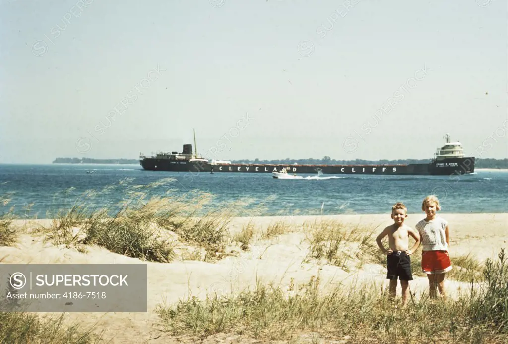 1950S Young Boy And Girl On Beach Smiling With A Tanker Ship On Lake Behind Them Port Huron Michigan 1955