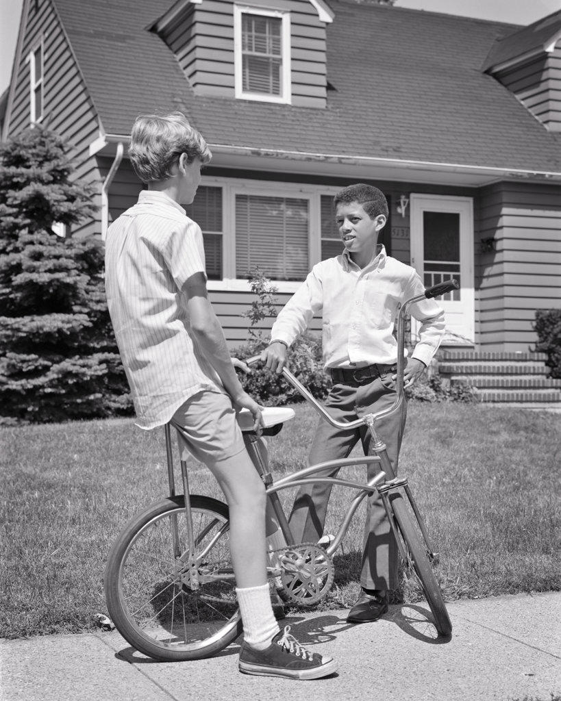 1960s 1970s TWO PRETEEN BOYS ONE AFRICAN AMERICAN AND THE OTHER CAUCASIAN WITH BICYCLE TALKING TOGETHER ON SUBURBAN SIDEWALK