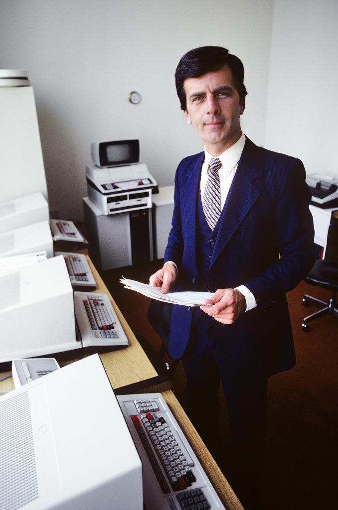 1980s MAN COMPUTER SYSTEMS SALESMAN STANDING LOOKING AT CAMERA IN A ROOM WITH EARLY HARDWARE MONITORS AND KEYBOARDS
