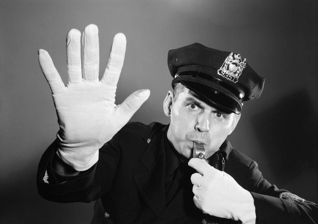 1950s 1960s POLICE MAN LOOKING AT CAMERA HAT WITH BADGE BLOWING WHISTLE WHITE GLOVED HAND UP TO HALT STOP TRAFFIC