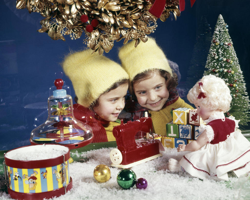 1960s TWO SMILING GIRLS TWIN SISTERS LOOKING IN CHRISTMAS TOY STORE WINDOW AT DOLL AND SEWING MACHINE