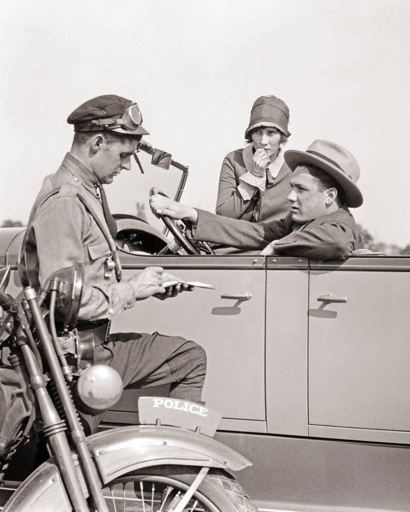 1920s MOTORCYCLE POLICEMAN WRITING A SPEEDING TICKET TO A COUPLE SITTING IN CONVERTIBLE SEDAN